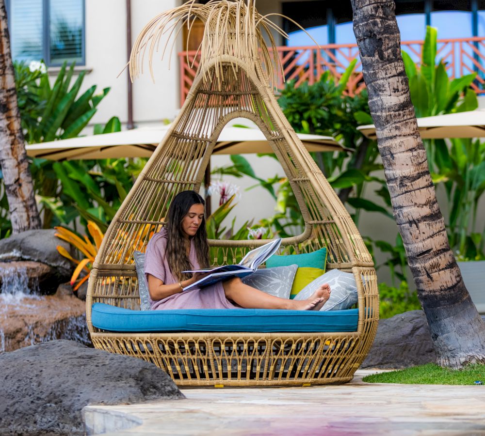 A woman sits reading in a stylish, teardrop-shaped wicker chair with cushions outside near tropical plants and trees, likely in a resort setting.