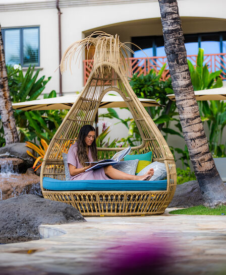 A woman sits reading in a stylish, teardrop-shaped wicker chair with cushions outside near tropical plants and trees, likely in a resort setting.