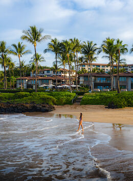 A person walks along a sandy beach with palm trees and buildings in the background, under a blue sky with some clouds.