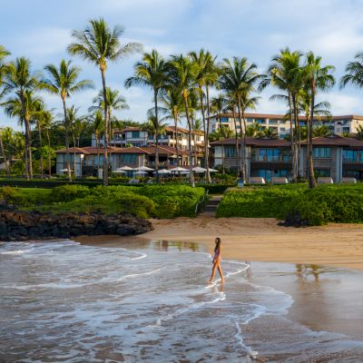 A person walks along a sandy beach with palm trees and buildings in the background, under a blue sky with some clouds.