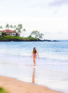 A woman walks along a sandy beach near the ocean with buildings and palm trees in the background, capturing a serene coastal scene at sunrise.