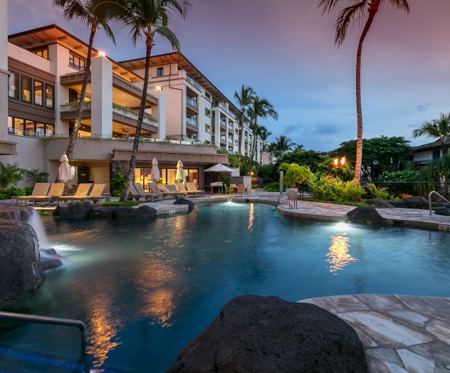 A serene resort pool area at dusk, featuring lounge chairs, a waterfall, palm trees, and a multi-story building in the background.