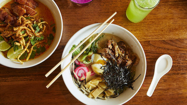 The image shows two bowls of Asian noodle dishes, chopsticks resting on one bowl, accompanied by two different beverages on a wooden table.