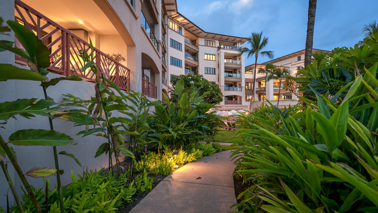 A pathway through lush greenery leads to a multi-story building with balconies, highlighted by evening lighting and a visible moon.