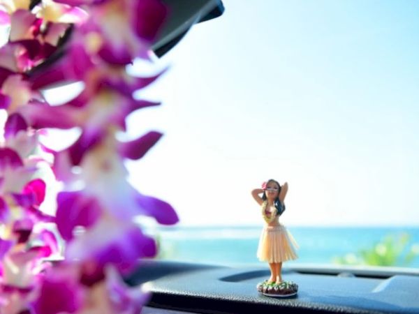 A hula girl figurine on a car dashboard with a blurry purple lei in the foreground and the ocean in the background, under a clear sky.