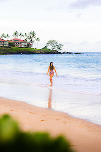 A person walks along a sandy beach, with waves lapping at their feet and palm trees in the background, creating a serene scene.