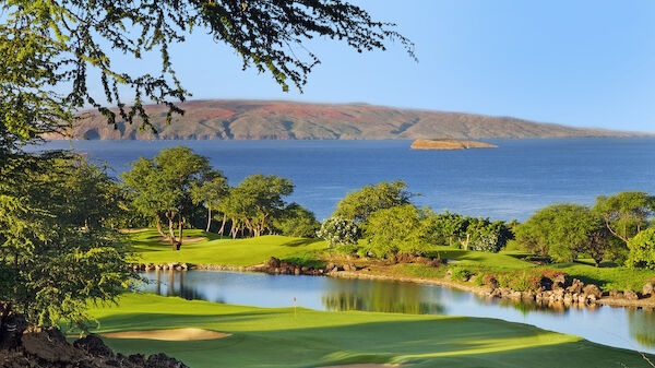 The image shows a scenic golf course with lush green fairways and water features, set against a backdrop of a calm blue ocean and an island in the distance.