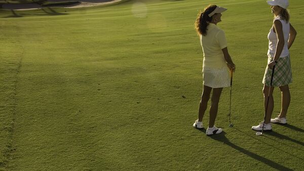 Two women are standing on a golf course, holding clubs, and conversing. The setting is sunny with shadows indicating late afternoon.