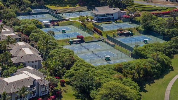 The image shows an aerial view of multiple tennis courts surrounded by greenery and buildings. The courts are separated by fences.