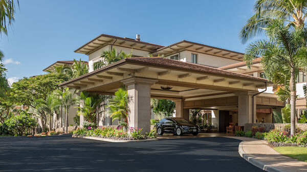 An elegant hotel entrance with a covered driveway, tropical landscaping, and luxury cars parked near the entrance under a clear blue sky.