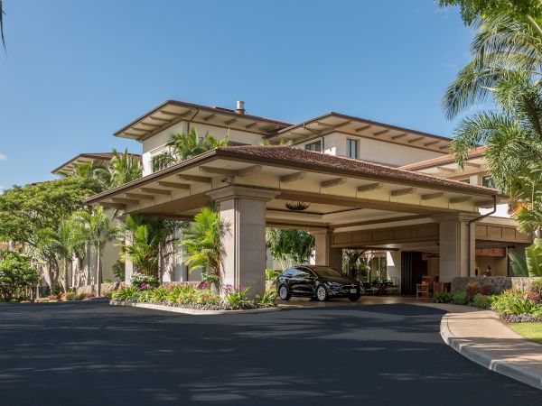 An elegant hotel entrance with a covered driveway, tropical landscaping, and luxury cars parked near the entrance under a clear blue sky.