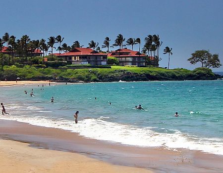 A scenic beach with white sand, turquoise waters, people swimming, and a backdrop of lush greenery along with red-roofed buildings.