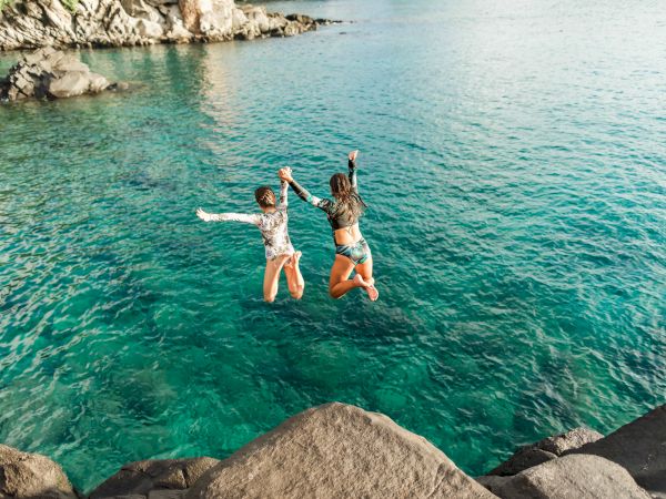 Two people jumping off rocks into clear blue water, holding hands, with a scenic rocky shoreline in the background.